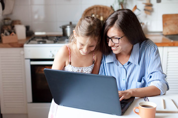 Holiday shopping online. Happy mother and child girl make purchases in the Internet on Black Friday. Cute kid and woman are smiling. Family are enjoying buying gifts with laptop in cozy home kitchen.