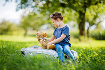 Wall Mural - Little preschool kid girl driving big vintage old toy car and having fun with playing with big plush toy bear, outdoors. Child enjoying warm summer day in nature landscape. Girl driving car