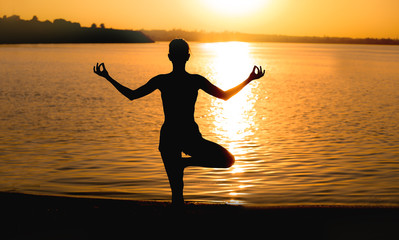 Wall Mural - Beautiful young woman practicing yoga near river at sunset