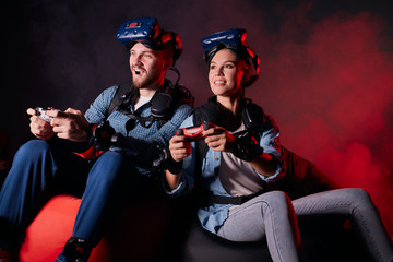 Wall Mural - Cheerful couple playing videogame sitting on sofa isolated over black background, neon backlit. Using joysticks, wearing headsets, goggles