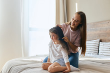 Pretty Charming Girl With Long Hair And Broad Smile Holding Small Hairbrush And Combing Brunette Hair Of Little Daughter Sitting On Bed, Morning Time Concept, Portrait