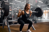 Charming strong female powerlifter dressed in black sportswear and white sneakers, doing squats, trying to stand with heavy barbell, professional sport concept, indoor shot