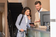 © Zoran Zeremski - Young businesswoman check-in in hotel, smiling male receptionist behind the hotel counter showing available rooms on tablet.
