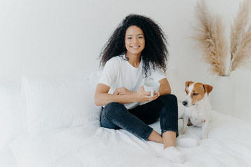 Wall Mural - Glad dark skinned Afro American female feels relaxed, poses in bedroom on comfortable bed with pedigree dog, drinks hot beverage, has morning coffee, smiles happily, enjoys spare time at home.