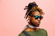 Close up portrait of a young man in green with dreadlocks and blue sunglasses, isolated on pink