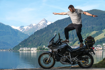 Biker man stand on top of a motorcycle keeps balance, extreme. hobby concept, adventure on two wheels. Sunny summer day in the Alpine mountains. Zell am see lake on background Austria.