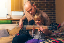 Cute Little Girl And Her Father Are Playing Bass Guitar And Smiling While Sitting On Couch At Home. Spending Time Together, Learning To Play The Guitar