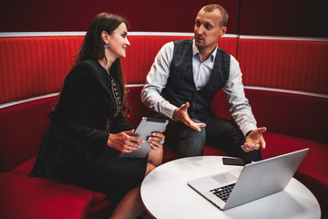 Business meeting of two colleagues with selective focus on a caucasian woman entrepreneur with a digital tablet speaking with his male partner; a laptop and smartphone on a table, bent red background