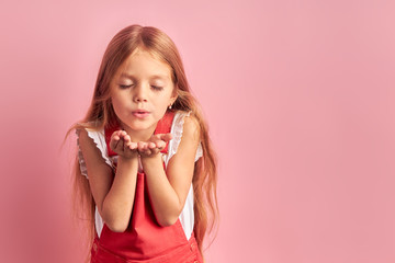 Portrait of little caucasian girl with long hair and closed eyes sending, blowing kiss to camera, wearing red overalls isolated over pink background.