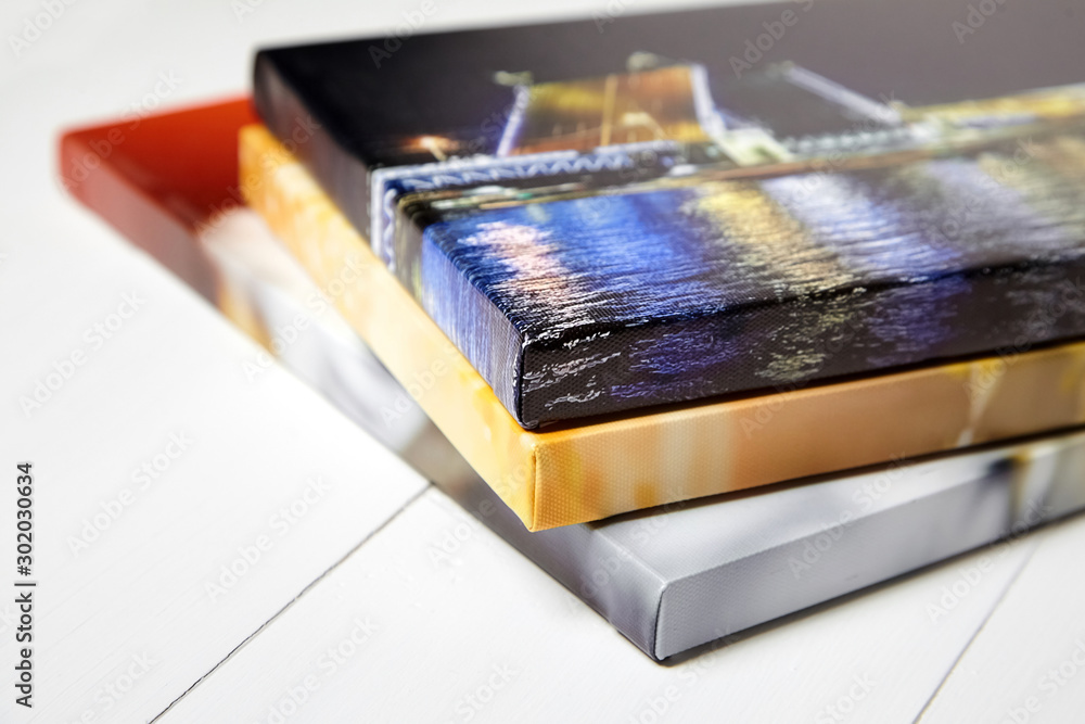 Photography canvas prints. Stack of colorful photos with gallery wrap on white wooden table. Stretched photo canvases, lateral side, closeup - obrazy, fototapety, plakaty 