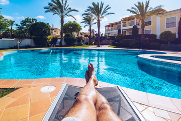 Relaxing at the swimming pool. Man relaxing next to swimming pool. Man enjoying the hot summer at swimming-pool. Sunbathing by the swimming pool, mans legs lying down on a sun lounger over the water.