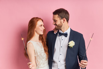 Portrait of young caucasian couple wearing dress and tuxedo holding sparklers isolated over pink background