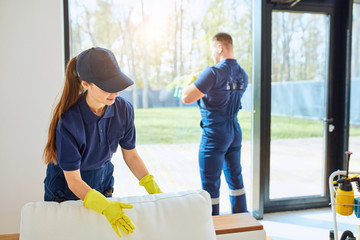 Caucasian janitors in blue uniform washing panoramic window, wiping dust off from sofa.