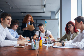 Wall Mural - Young colleagues, caucasian business partners working as designers sit together at table in modern office with panoramic window. Papers, colorful pencils on table, active discussion