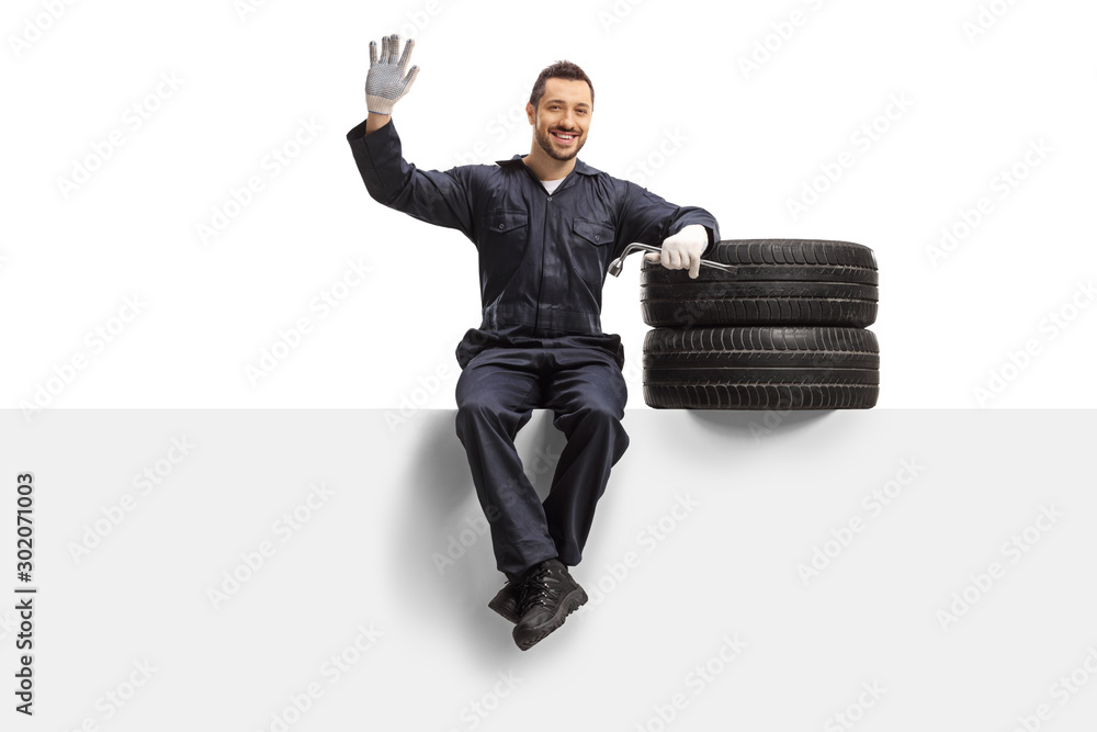 Auto mechanic sitting on a panel with tires and a wrench and waving at the camera - obrazy, fototapety, plakaty 