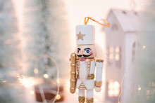 Christmas Nutcracker Toy Soldier Figurine Ornament In White. Decoration For New Year.  Nutcracker On The White Sparkling Background With Conifers. Advent Concept With Bokeh Lights.