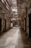 Fototapeta Uliczki - Hallway of an old abandoned prison showing doorways to cells, metal stairs, and bars on the ceiling.
