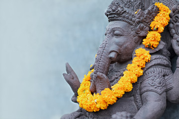 Ganesha sitting in meditating yoga pose in front of hindu temple. Decorated for religious festival by orange flowers garland, ceremonial offering. Balinese travel background. Bali island art, culture.