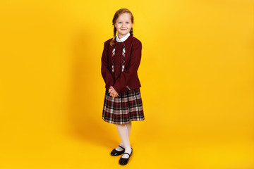 A full-length schoolgirl stands and looks at the camera. Cute little girl in uniform on a yellow background.