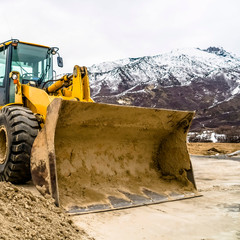 Wall Mural - Square frame Front view of a yellow bulldozer against snow topped mountain and cloudy sky