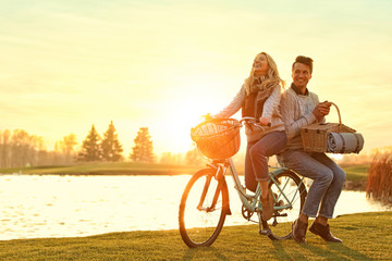 Young couple with bicycle and picnic basket near lake on sunny day