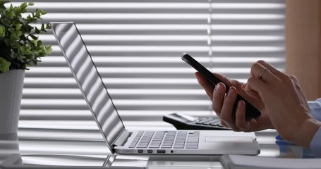 Person using smartphone and laptop computer in office for financial analysis or business management, working with internet, horizontal dolly shot, 4k video footage