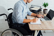 © LIGHTFIELD STUDIOS - Cropped view of man in wheelchair writing in notebook and using laptop at workplace
