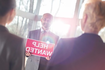 Wall Mural - Smiling senior businessman showing help wanted sign to businesswomen while standing at office