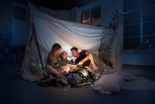 Caucasian Family Sitting In A Teepee, Reading Stories With The Flashlight In Dark Room With Toys And Pillows. Caucasian Models. Home Comfort, Family, Love, Christmas Holidays, Storytelling Time.