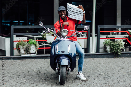 Friendly african guy holds cardboard boxes, one opened container with delicious pizza, transports food items from cafe to clients, brings orders by motorbike, likes his job.