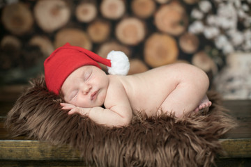 Christmas newborn baby boy girl sleeping in front of a pile of wood logs in a cabin with a red santa claus hat on a rug