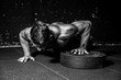© Srdjan - Young strong sweaty focused fit muscular man with big muscles performing push ups with one hand on the barbell weight plate for cross training hard core workout in the gym  black and white