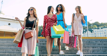 Wall Mural - Walking in the city. A group of four splendid girls is walking along the urban streets after the shopping, wearing bright outfits, chatting and laughing.