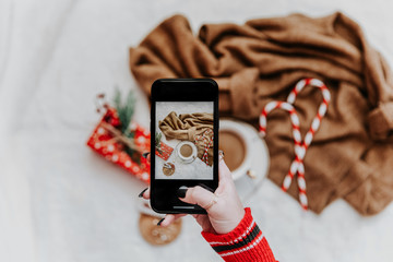 Woman's hand take a mobile photo of christmas composition with hot coffee. Smartphone with mobile camera display while shooting. Top view