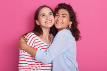 Two Pretty Sisters Or Friends Women With Dark Hair, Wearing Casual Shirts. Girls Hugging Over Pink Background In Studio, Ladies Having Happy Expression, Celebrating Something, Look With Charming Smile
