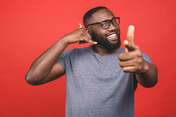 Young african american man wearing casual t-shirt standing over isolated red background smiling doing talking on the telephone gesture and pointing to you. Call me.