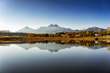 Wall Mural - Peaceful scene of beautiful autumn mountain landscape with small settlement, calm lake, colorful trees and high peaks in High Tatras, Slovakia.