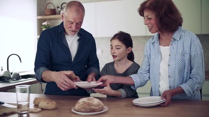 Sticker - A small girl with senior grandparents indoors in kitchen preparing food.