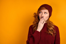 A Curly Red-haired Girl In A Burgundy Sweater And Hat Stands On An Orange Background, Looks Sideways, Covering Her Mouth With Palm