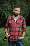 Fototapeta Młodzieżowe - Bearded hipster in red shirt and dark blue jeans is hoolding a bottle of wine and looking at the camera. Bad habits concept