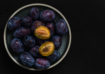 Ceramic plate with ripe plums over a dark background. View from above. Prunes with water droplets, two slices of cut prunes, space for text. Agriculture and gardening, fresh harvest, concept.