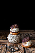 Homemade Choux Pastries With Cream And Chocolate Scissors And Ball Of Yarn Arranged On Texture Wooden Surface Against Black Background
