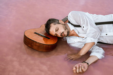 Sleepy Man In White Shirt And Suspenders Lying Down With Closed Eyes With Head Resting At An Acoustic Guitar In Sea At Sandbank