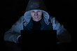 A criminal in the hood sits in the dark in front of a laptop monitor, cybercrime