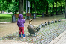 Unrecognizable Little Girl With Her Back To Us Counts The Number Of Little Ducklings Of Mothers Duck, Sculpture In The Park