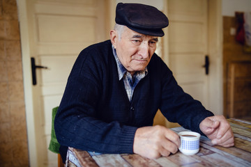 Senior man grandfather old pensioner farmer wearing black sweater and hat having a cup of coffee or tea by the table at home sitting alone