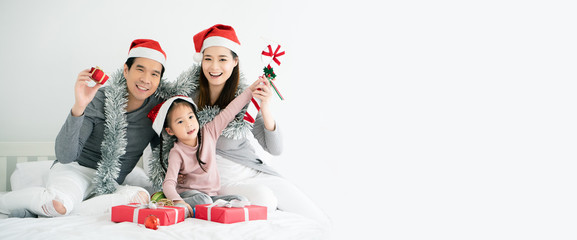 Asian family have father, mother and daughter with gray, pink casual wear and Santa hats. Sitting on a white bed with a red gift box And  small ball. Celebrate Christmas and Happy New Year. copy space