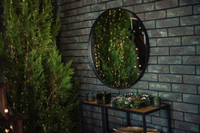 On A Khaki Brick Green Wall Hangs A Round Mirror With A Black Frame. Around The Christmas Trees In Garlands With Lights, Juniper. Wallpaper For Advertising, Text, Blurred Background. Copy Space Place