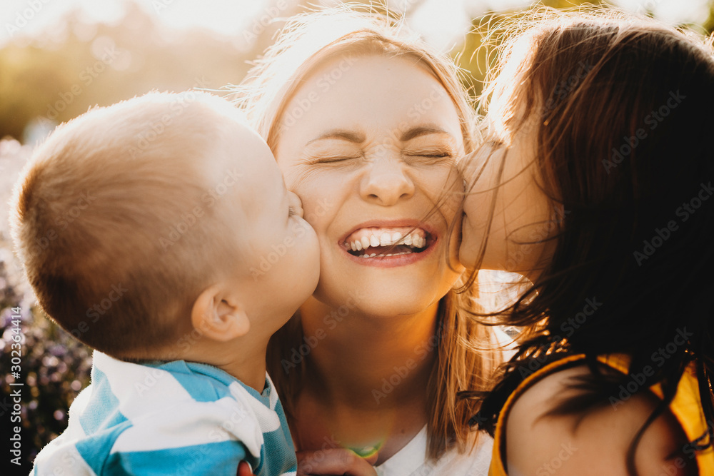 Close up portrait of lovely young mother laughing with closed eyes while her kids is kissing her on the cheeks outdoor against sunset. - obrazy, fototapety, plakaty 