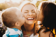 Close Up Portrait Of Lovely Young Mother Laughing With Closed Eyes While Her Kids Is Kissing Her On The Cheeks Outdoor Against Sunset.